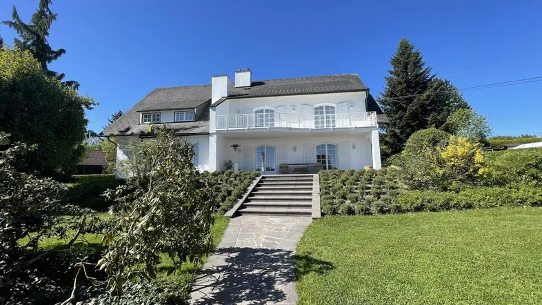 Elegantes weißes Haus mit gepflegtem Vorgarten, Steintreppe und Balkon unter blauem Himmel.