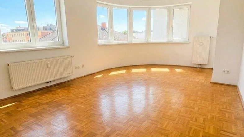 Helles Zimmer mit Parkettboden und mehreren Fenstern, die viel Tageslicht hereinlassen.