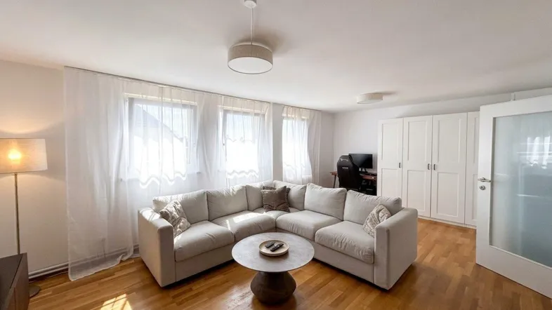Helles Wohnzimmer mit beigem Ecksofa, Parkettboden, großen Fenstern und weißem Einbauschrank.