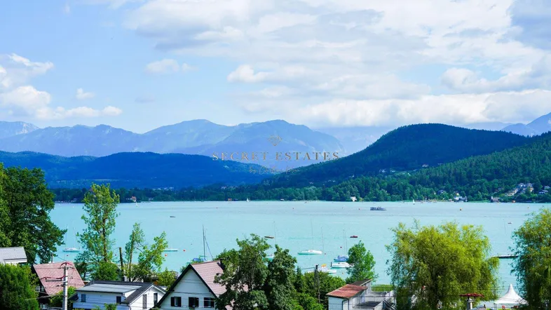 Panoramablick auf den See mit umliegenden Häusern, grünen Hügeln und majestätischen Bergen im Hintergrund.