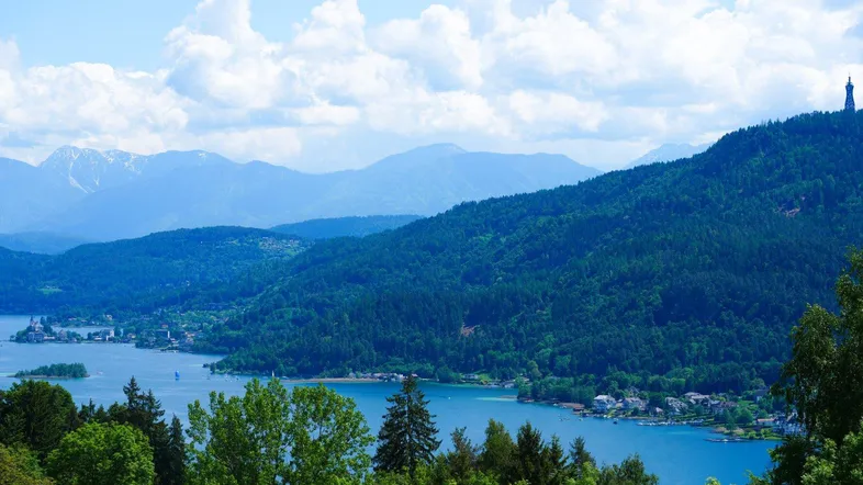 Weitläufige Landschaftsaufnahme mit einem glitzernden See, dichten Wäldern und einer beeindruckenden Bergkette im Hintergrund.