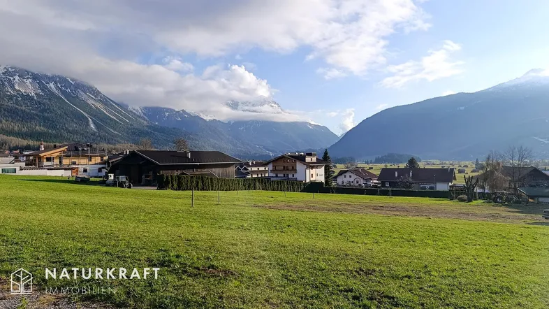 Ländliche Umgebung mit Häusern, grünen Wiesen und majestätischen Bergen im Hintergrund unter bewölktem Himmel.
