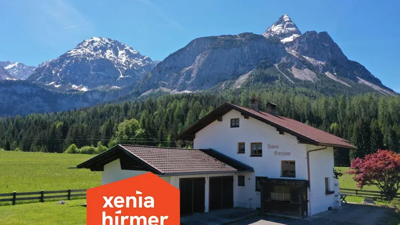 Traditionelles Haus mit Satteldach und Garage, umgeben von grünen Wiesen und majestätischen Bergen.