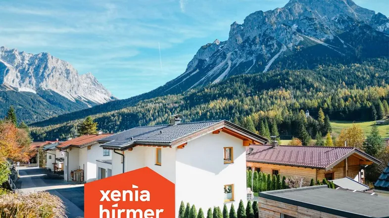 Weißes Haus mit dunklem Dach in alpiner Umgebung, umgeben von Bergen und Bäumen.