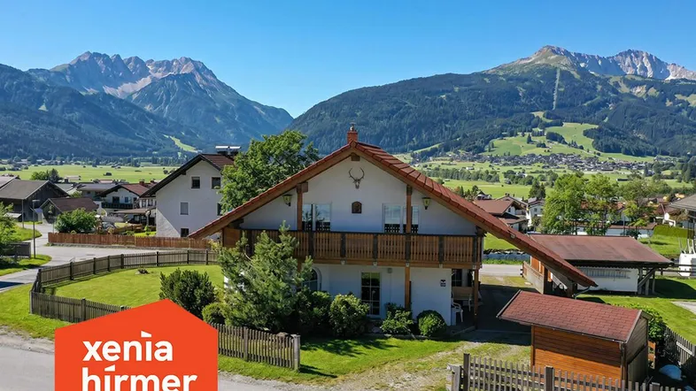 Traditionelles Haus im alpinen Stil mit Balkon und Bergblick, umgeben von grüner Landschaft.