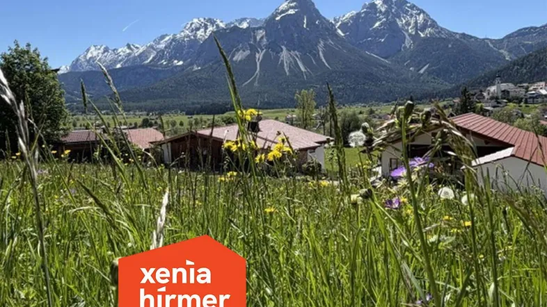Panoramablick auf Häuser in einer grünen Wiese mit majestätischen Bergen im Hintergrund und Immobilienlogo.