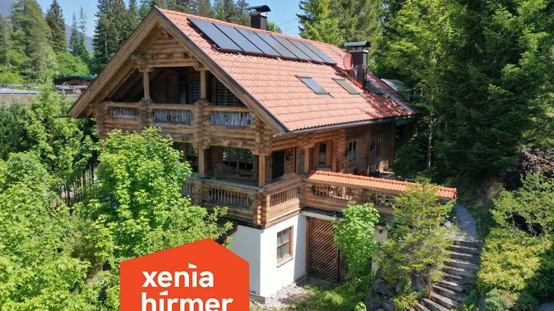 Traditionelles Holzhaus im alpinen Stil mit Ziegeldach und Solarpaneelen, umgeben von üppiger Natur.