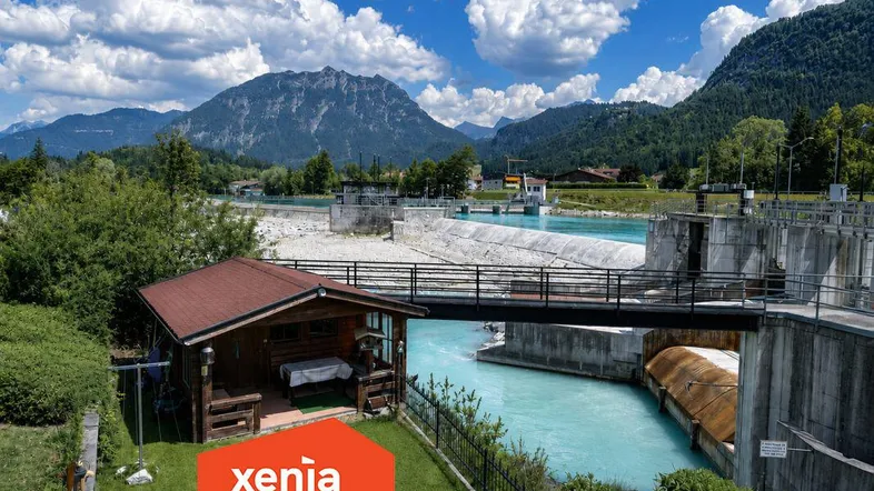 Idyllisches Anwesen am Fluss mit Bergblick und kleinem Holzhaus unter blauem Himmel.