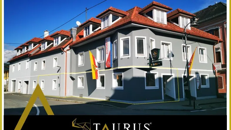 Graues Gebäude mit rotem Ziegeldach und mehreren Dachgauben an einer Straßenecke, mit Werbebanner und Logo.