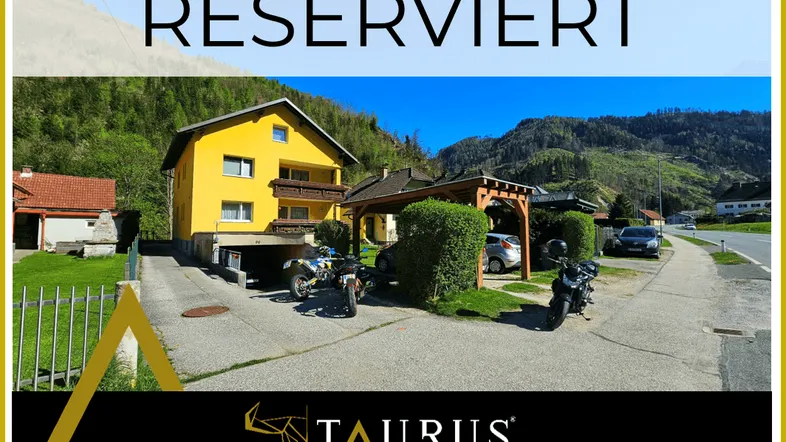 Gelbes Wohnhaus mit Carport und Stellplätzen in einer malerischen Berglandschaft.