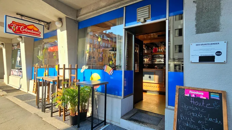 Eingangsbereich eines kolumbianischen Restaurants mit blauer Fassade und Außentischen an einem sonnigen Tag.