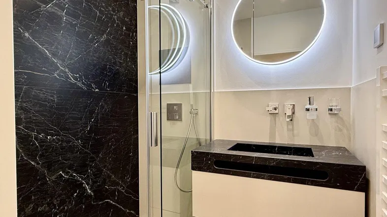Modernes Badezimmer mit dunklen Marmorfliesen in der Dusche und beleuchtetem Spiegel.