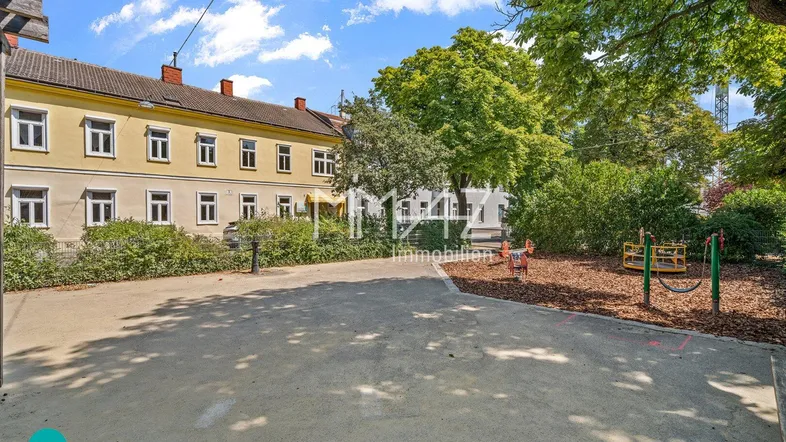 Gelbes Mehrfamilienhaus mit einem kleinen Spielplatz im Vordergrund unter blauem Himmel.