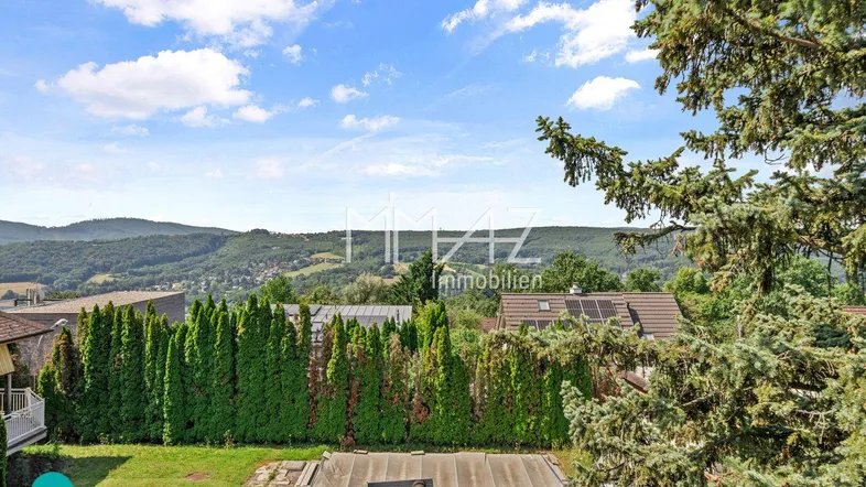 Blick auf die umliegende Hügellandschaft mit viel Grün und einem Teil des Gartens unter blauem Himmel.