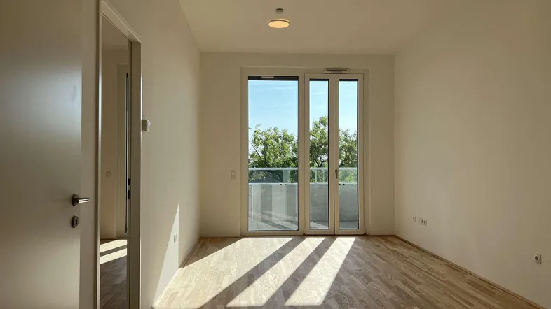 Helles, leeres Zimmer mit Holzboden und Zugang zu einem Balkon, durchflutet von Sonnenlicht.