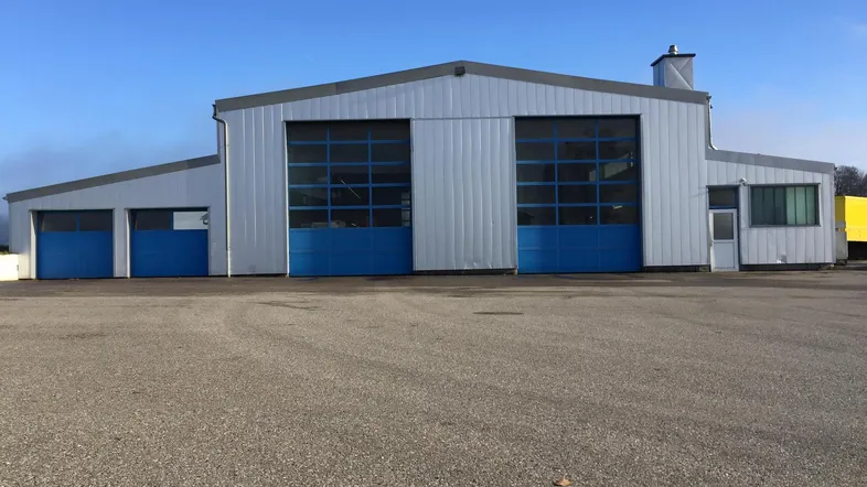 Ansicht des Gewerbegebäudes mit großen blauen Rolltoren und einem weitläufigen Vorplatz unter blauem Himmel.