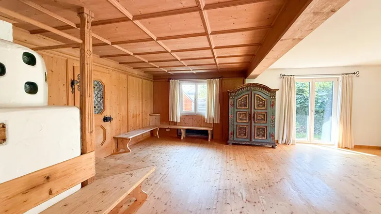 Traditionelles Wohnzimmer mit Holzboden, Holzwänden, Holzbalkendecke und einem Kachelofen.
