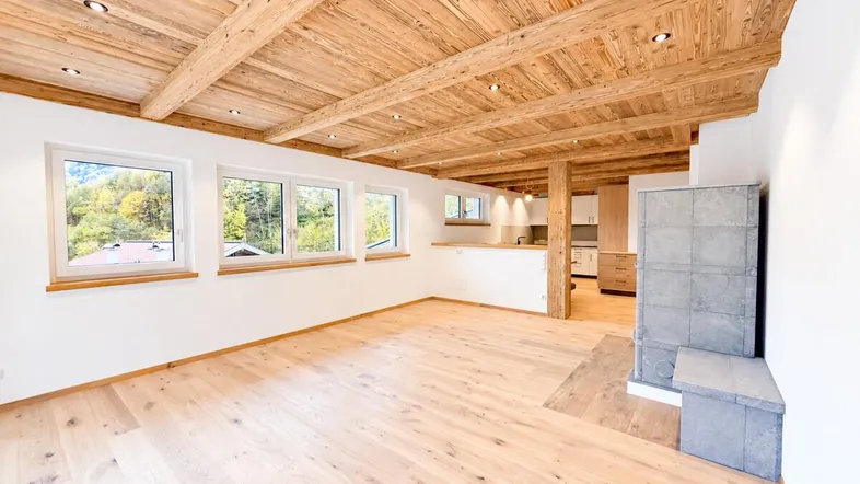 Helles Wohnzimmer mit Holzbalkendecke, Parkettboden und modernem Kaminofen, offen zur Küche.