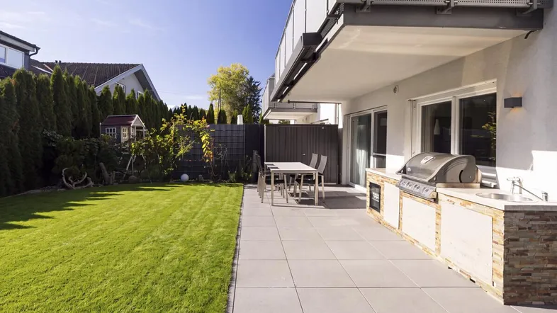 Große gepflasterte Terrasse mit Outdoor-Küche und angrenzendem Rasenbereich, ideal für Entspannung.