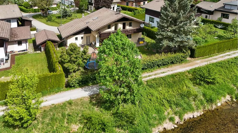 Luftaufnahme eines Einfamilienhauses mit Garten, umgeben von viel Grün und einem Bachlauf.