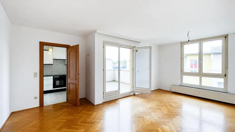 Helles Wohnzimmer mit Fischgrätparkett, Zugang zum Balkon und Blick in die angrenzende Küche.