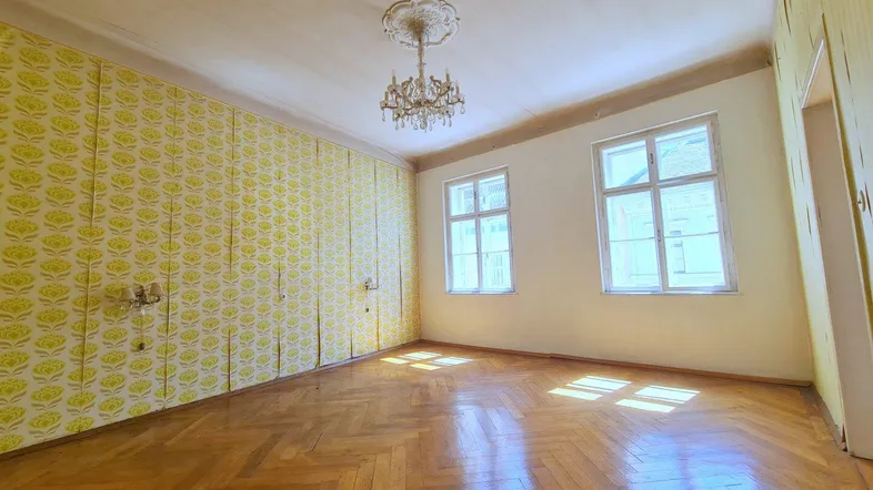 Helles Zimmer mit Fischgrätparkett, Kronleuchter und hohen Decken, bereit zur Renovierung.
