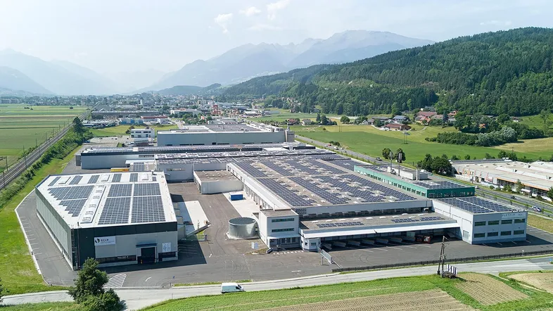 Luftaufnahme eines großen Industrie- und Gewerbekomplexes mit zahlreichen Solarmodulen auf den Dächern, umgeben von grünen Feldern und Bergen.
