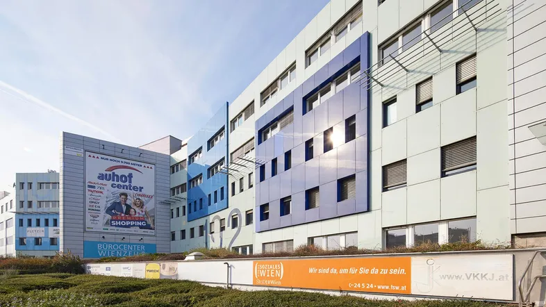 Modernes Bürogebäude mit blauen und hellgrauen Fassadenelementen unter blauem Himmel und umliegender Bepflanzung.