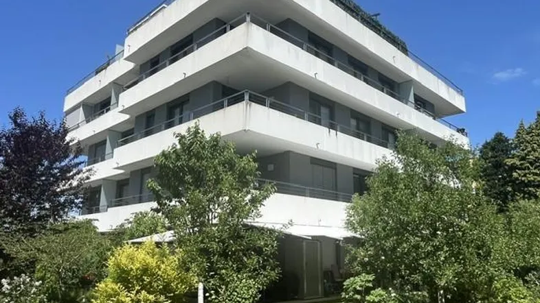 Modernes Mehrfamilienhaus mit mehreren Balkonen und begrüntem Vorgarten unter blauem Himmel.