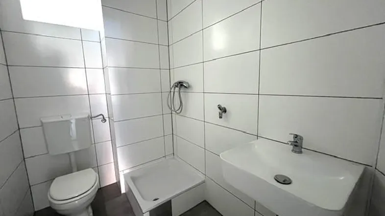 Helles Badezimmer mit weißen Fliesen, Dusche, WC und modernem Einzelwaschbecken.