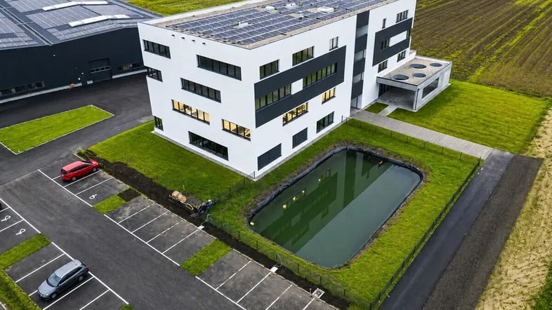 Modernes Geschäftsgebäude mit Solaranlage, Teich und Parkplätzen, umgeben von Grünflächen und weiteren Gewerbebauten.