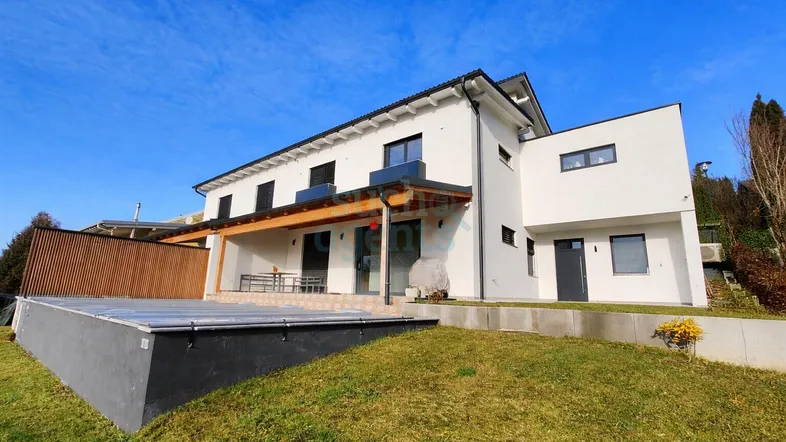 Modernes Einfamilienhaus mit weißer Fassade, großem Garten und überdachtem Poolbereich unter blauem Himmel.