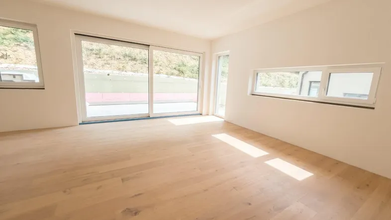 Helles, leeres Zimmer mit Holzboden und großen Schiebetüren, die auf eine Terrasse führen.