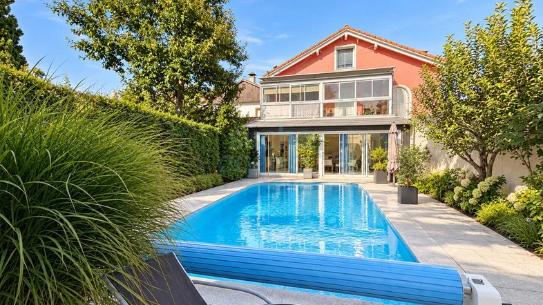 Großer Außenpool mit blauer Abdeckung und Liegestuhl, umgeben von gepflegtem Garten und Wohnhaus.