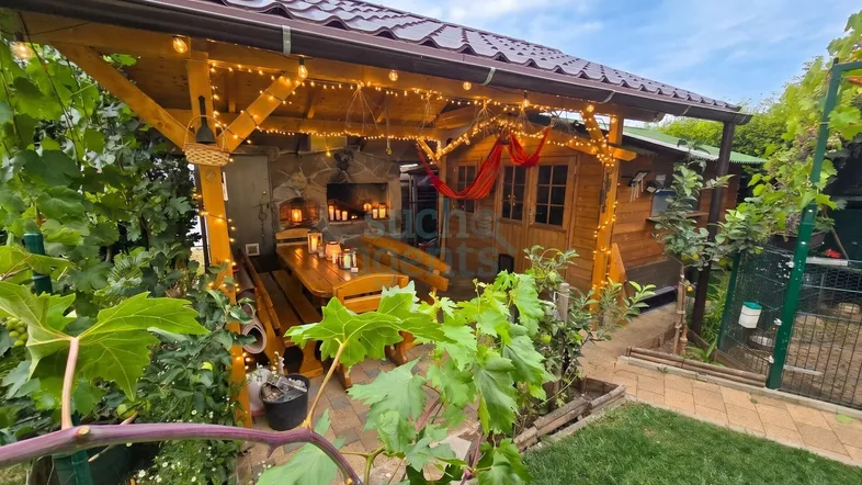 Überdachte Terrasse mit Holztisch, Bänken, Lichterketten und integriertem Grillbereich, umgeben von Grün.