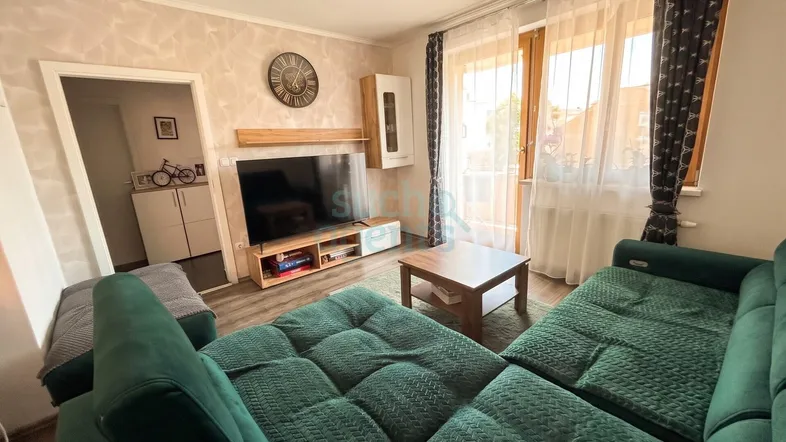 Helles Wohnzimmer mit großem Sofa, Fernseher und Zugang zum Balkon.