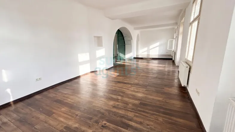 Geräumiger, heller Raum mit dunklem Holzfußboden, weißen Wänden und mehreren Fenstern, die viel Tageslicht hereinlassen.