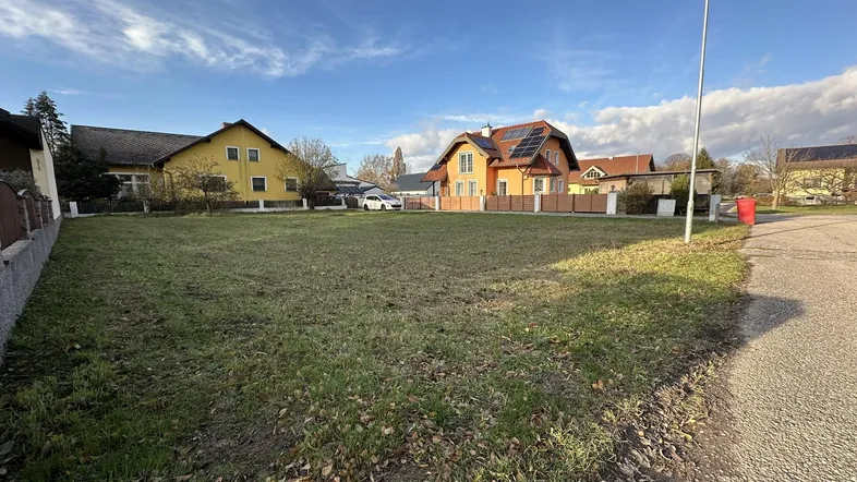 Leeres Grundstück mit angrenzender Straße und umliegenden Wohnhäusern unter blauem Himmel.