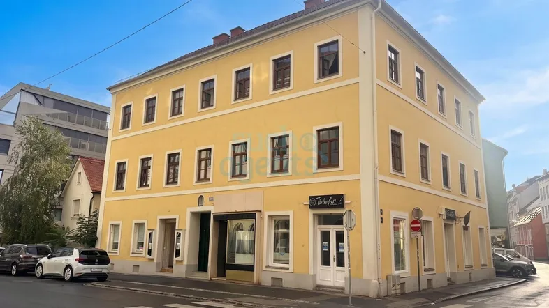 Ein gut erhaltenes Eckgebäude mit gelber Fassade, das im Erdgeschoss Gewerbeflächen und in den oberen Etagen Wohnungen beherbergt.
