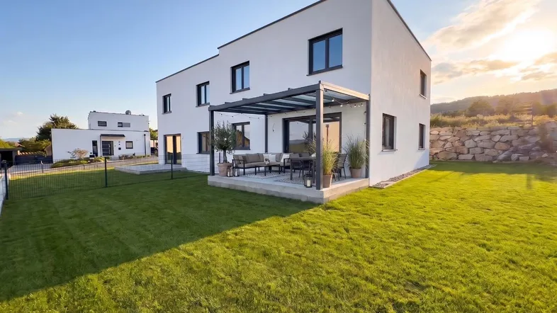 Modernes weißes Haus mit großer Rasenfläche, überdachter Terrasse und Blick ins Grüne.