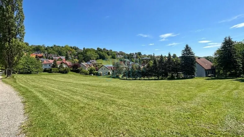 Weitläufige Grünfläche mit Blick auf eine Siedlung am Hügel unter blauem Himmel.