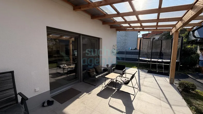 Sonnige Terrasse mit moderner Überdachung, Sitzgelegenheiten und Zugang zum Garten mit Trampolin.