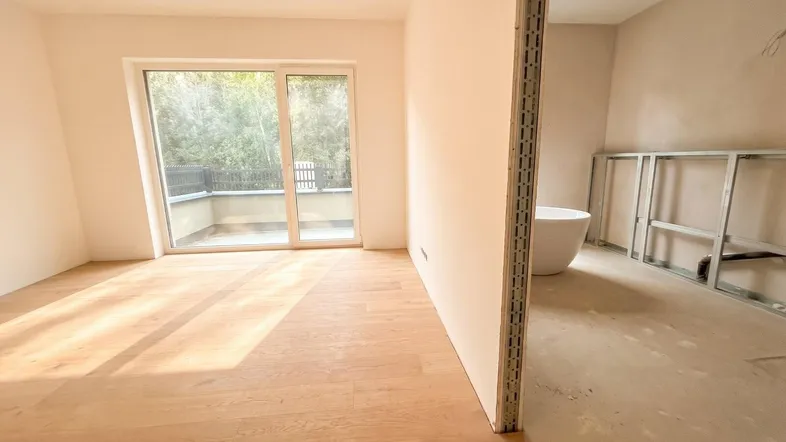 Helles, unfertiges Zimmer mit Holzboden und Zugang zu einem Balkon, angrenzend an ein Badezimmer im Bau.