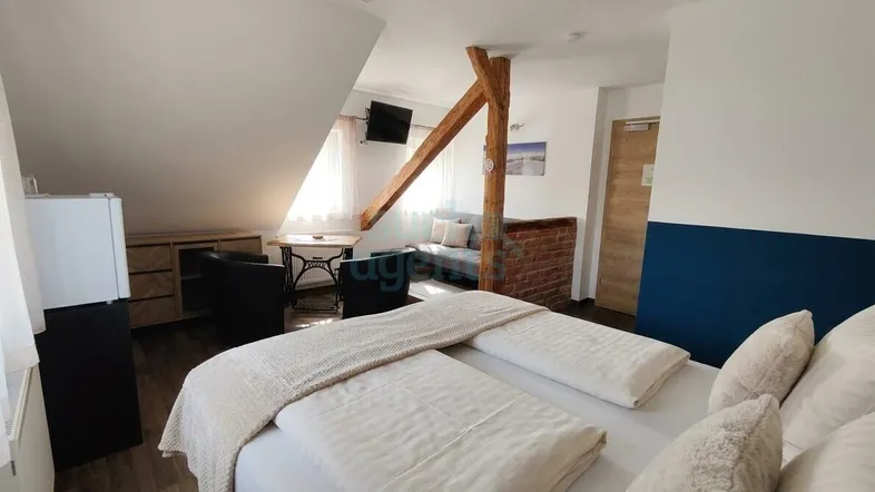 Helles Schlafzimmer im Dachgeschoss mit schräger Decke, Holzbalken und einem kleinen Sitzbereich.