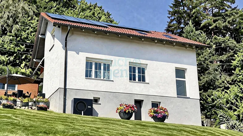 Modernes Einfamilienhaus mit weißer Fassade, rotem Ziegeldach und Photovoltaikanlage auf einem grünen Hügel.