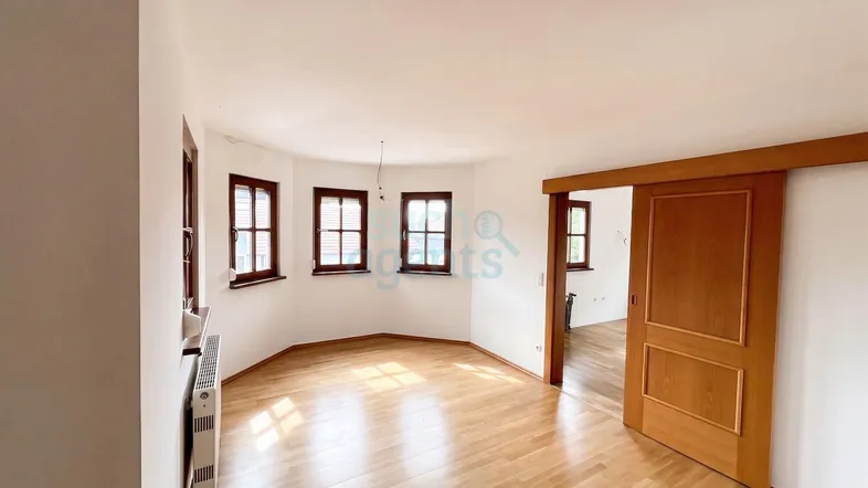 Heller Wohnraum mit Parkettboden, mehreren Fenstern und einer Holzschiebetür zu einem angrenzenden Zimmer.