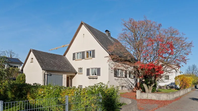 Hausfassade mit Satteldach, Fensterläden und einem Baum mit Herbstlaub unter blauem Himmel.