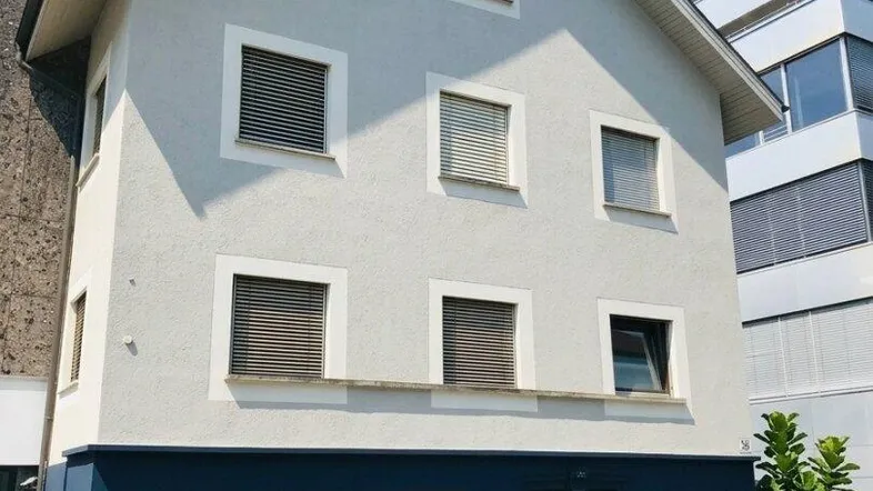 Gut erhaltenes Gebäude mit grauer Fassade, blauem Sockel und Rollläden unter strahlend blauem Himmel.