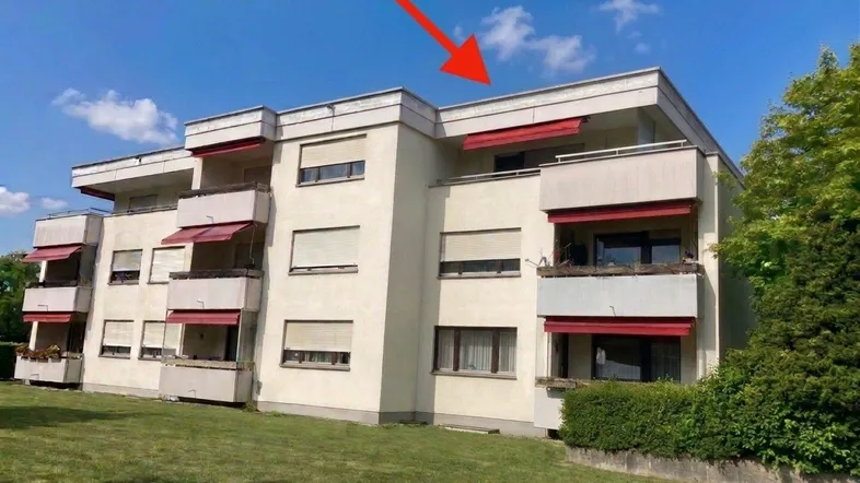 Mehrfamilienhaus mit Balkonen und roten Markisen unter blauem Himmel und grüner Wiese.
