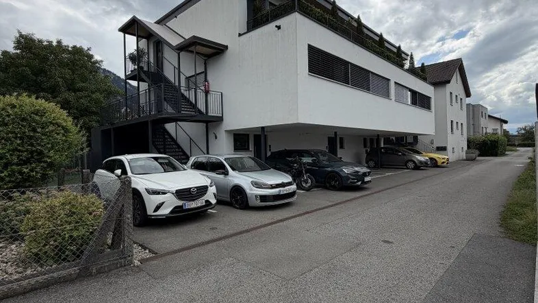 Modernes Mehrfamilienhaus mit weißer Fassade, Balkonen und Parkplätzen im Freien unter bewölktem Himmel.